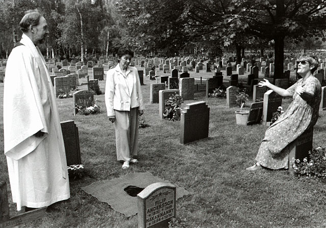 Two sisters by the urn grave of their mother. A priest in white gown standing by