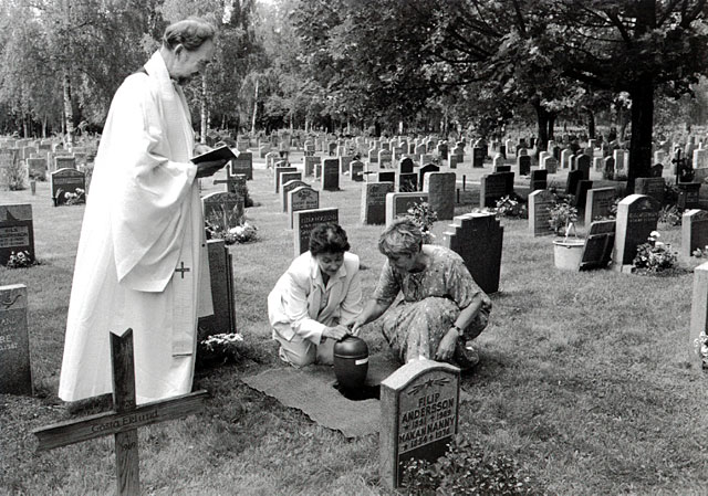 Interment of the urn. Two sisters lowering the urn of their mother. A priset in white gown standing by