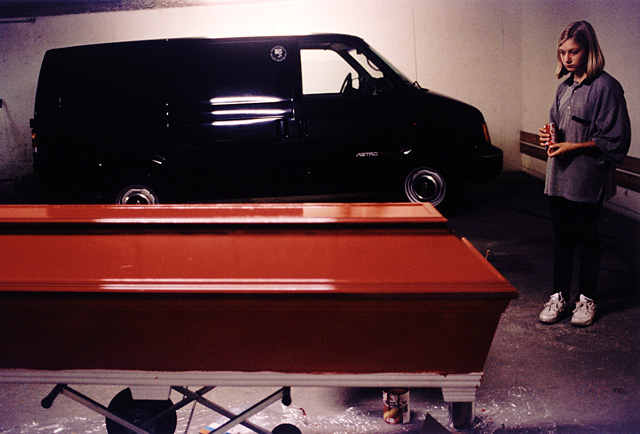 Granddaughter with Coca Cola can in her hands watching the red coffin, black herse in the background