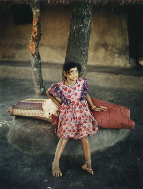 Girl with skirt with flowers sitting underneath a palm tree on her front yard, Kudle India