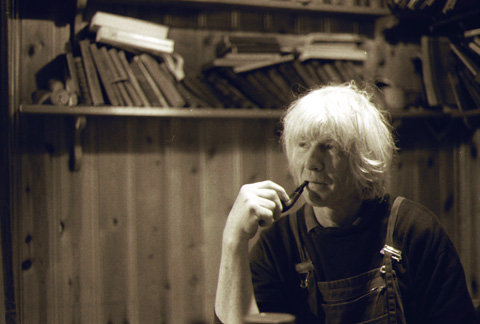 Man with white hair smoking pipe with books in background