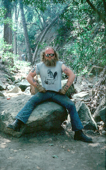 Scary looking motorcycle guy with beard sun glasses, tattoos and a T-shirt with a man sitting on toilet