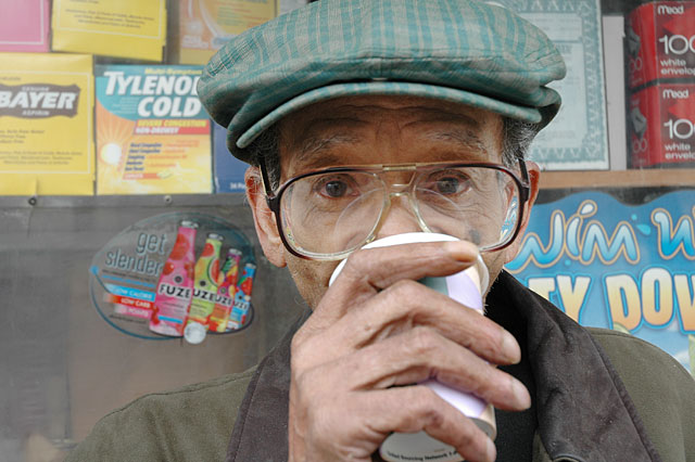 Man with big eye glasses and a green hat drinking coffee